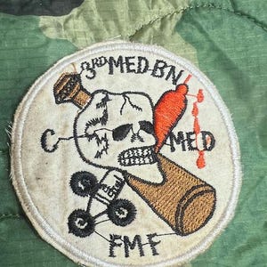 May include: Embroidered patch with a skull, bottle, and medical imagery. The patch features the text "3rd MED BN" and "FMF" in black lettering. The design includes a brown bottle, a red medical symbol, and a skull with crossbones.