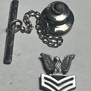 May include: A silver-toned tie tack set featuring a round head, a chain, and a bar. Includes a separate silver-toned emblem of an eagle with outstretched wings above a chevron design.