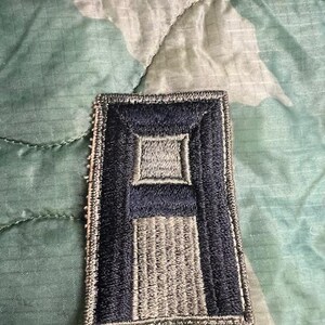 May include: A black and gray embroidered patch with the letter "A" in the center. The patch is surrounded by a black border and has a gray background.