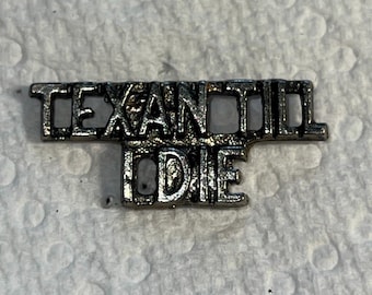 Op de afbeelding: Een zilverkleurige metalen pin met de woorden "TEXAN OIL DIE" in hoofdletters. De pin heeft een verweerde, vintage uitstraling, met een licht aangetaste afwerking. De letters zijn blokkerig en vet.