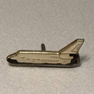 May include: A gold-colored pin shaped like a space shuttle. The shuttle is white with black details.