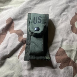 May include: A green canvas pouch with a flap secured by a black snap. The letters "US" are printed in black on the flap. The pouch is set against a camouflage background.