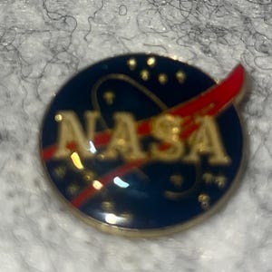 May include: A round NASA pin with a blue background and gold lettering. The NASA logo features a red swoosh and white stars. The pin has a gold border and is likely made of metal.