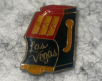 Op de afbeelding: Een zwarte, rode en gouden gokautomaatspeld met de woorden "Las Vegas" in gouden letters. De speld heeft drie gele rollen en een gouden hendel. De speld is een souvenir of verzamelobject.