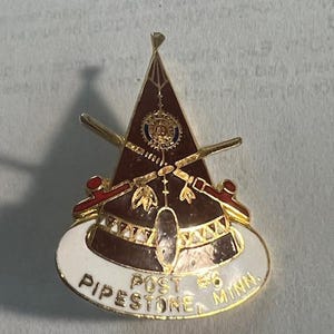 May include: A gold-colored pin with a white background. The pin features a brown teepee with a red and white design. The text "POST #6 PIPESTONE MINN" is written on the white background.