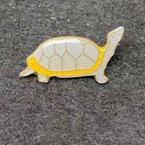 May include: A small, white turtle pin with a yellow shell. The turtle is facing right and has a detailed shell pattern.