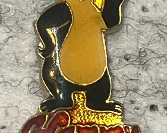Op de afbeelding: Een vintage speld met een cartoon panda beer die een biertje vasthoudt. De beer is zwart, goud en bruin, staand op een gouden basis met het woord "Hamm's" in rode en gouden letters. Een verzamelobject.