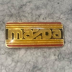 May include: Rectangular Mazda emblem with a gold-colored "Mazda" text. The emblem has a black border and a striped background in shades of red and gold. The emblem is on a gray, textured surface.