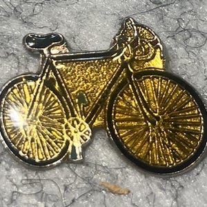 May include: A gold-colored bicycle pin with black accents. The pin features a detailed design of a bicycle with spokes and a seat. The bicycle is set against a textured background.