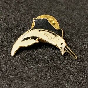 May include: A gold-tone pin featuring a black and white marlin fish design. The marlin is jumping out of the water with its dorsal fin raised.