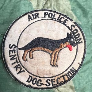 May include: A circular patch with the text "AIR POLICE SQDN" and "SENTRY DOG SECTION" encircling a black and tan dog illustration with a red tongue. The patch is on a camouflage background.