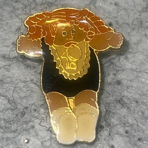May include: A gold-tone metal enamel pin featuring a stylized doll with brown hair in pigtails, wearing a black outfit with a light yellow bib that has a small animal graphic, and light-colored socks. The pin has a polished gold metal outline.