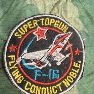 May include: A black and white embroidered patch with the text "SUPER TOPGUN FLYING CONDUCT NIBLE" in a yellow circle. A red star with white outline is in the upper left corner of the circle. A white and blue F-16 fighter jet is in the center of the circle.
