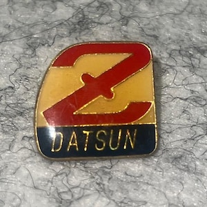 May include: A vintage Datsun pin featuring a red 'Z' emblem over a yellow background, with the word 'DATSUN' in white lettering on a navy blue base. The pin has a gold border.