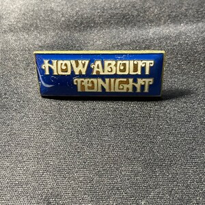 May include: A gold-colored pin with a blue background and the text "NOW ABOUT TONIGHT" in white.