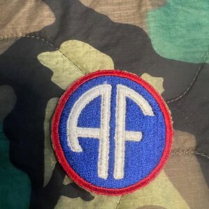 May include: A blue and red embroidered patch with the letters "AF" in white inside a circle. The patch is a military insignia.