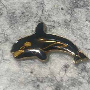 May include: A black and gold enamel pin in the shape of an orca whale. The pin features a glossy black body with gold accents on the fins, belly, and eye details. The pin is set against a textured, light gray background.