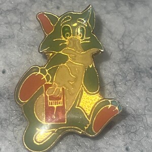 May include: A gold-toned metal enamel pin featuring a cartoon cat character with olive green fur, a tan chest, and reddish-brown accents on the ears and feet. The cat is depicted in a seated position, holding a small rectangular object with a red base.
