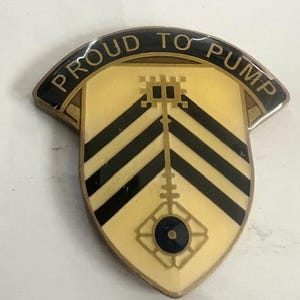 May include: A shield-shaped pin with the words "PROUD TO PUMP" in gold lettering on a black banner. The pin features a gold background with black stripes and a central design. The pin has a gold border.