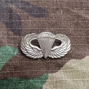 May include: A silver paratrooper badge with wings and a parachute, on a camouflage fabric background.