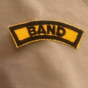 May include: A yellow and black fabric patch with the word "BAND" embroidered in black.