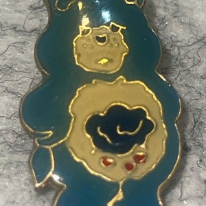 May include: A teal enamel pin shaped like a cartoon bear with a gold-toned metal outline. The bear features a cream-colored belly patch displaying a dark blue rain cloud graphic with three small red raindrops falling from the bottom of the cloud.