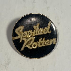 May include: A round, black pin with the words "Spoiled Rotten" in gold lettering. The pin has a gold border and a slightly worn appearance.