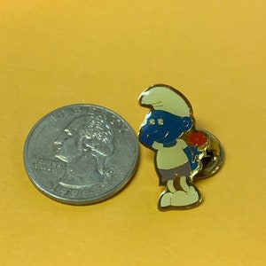 May include: A gold-colored pin featuring a cartoon character. The character is a blue Smurf with a white hat and a red flower. The Smurf is holding a red flower in its right hand. The pin is about the size of a quarter.