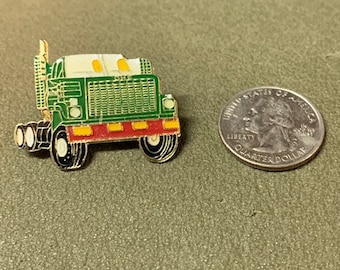 Op de afbeelding: Een groene en rode metalen pin van een vrachtwagen met een witte cabine. De pin is naast een Amerikaanse kwart dollar voor groottevergelijking.