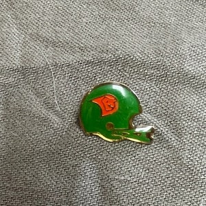 May include: Green and orange football helmet pin. The helmet is a stylized design with a small orange football inside.