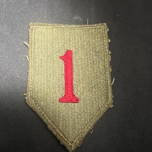 May include: A military patch with a shield shape, featuring a red number 1 on a green background. The patch has a textured surface and a stitched border. The patch is likely for a military uniform.