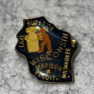 May include: A vintage Wisconsin state pin in the shape of the state, with a dark blue background and gold trim. The pin features a gold milk bottle and a figure, with text including "Superior", "Wisconsin", "Madison", and "Milwaukee".
