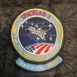 May include: A blue and yellow embroidered patch with the text "SPACELAB 3" and the names "LIND THAGARD OVERMYER GREGORY THORNTON VAN DEN BERG WANG". The patch features a space shuttle in orbit around the Earth.