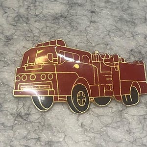 May include: A red and gold-toned fire truck pin. The pin features a detailed design of a fire truck with black wheels and gold trim. The pin is likely made of metal and has a glossy finish.