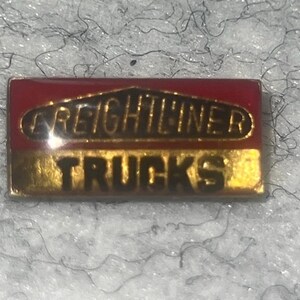 May include: A rectangular metal pin featuring a red top section with the word FREIGHTLINER in black letters inside a gold-bordered oval shape, and a gold bottom section with the word TRUCKS printed in bold black capital letters.