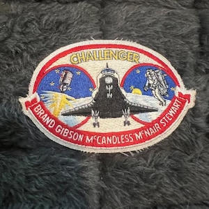 May include: A vintage embroidered patch featuring the Space Shuttle Challenger with the names of the crew members: Brand Gibson, Mc Candless, M'Nair, Stewart. The patch is red, white, and blue with a black and white image of the shuttle in space.