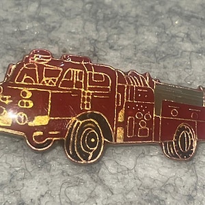 May include: A metallic enamel pin shaped like a fire engine, featuring a deep red body with gold-toned outlines and detailing. The design depicts the side profile of the emergency vehicle, including wheels, windows, and equipment compartments.