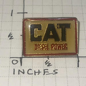 以下が含まれることがあります： 黒と赤の文字で「CAT DIESEL POWER」と書かれた長方形のピン。ピンは金色の背景に赤い縁取りがあります。ピンの長さは約1.9cm、幅は約1.3cmです。