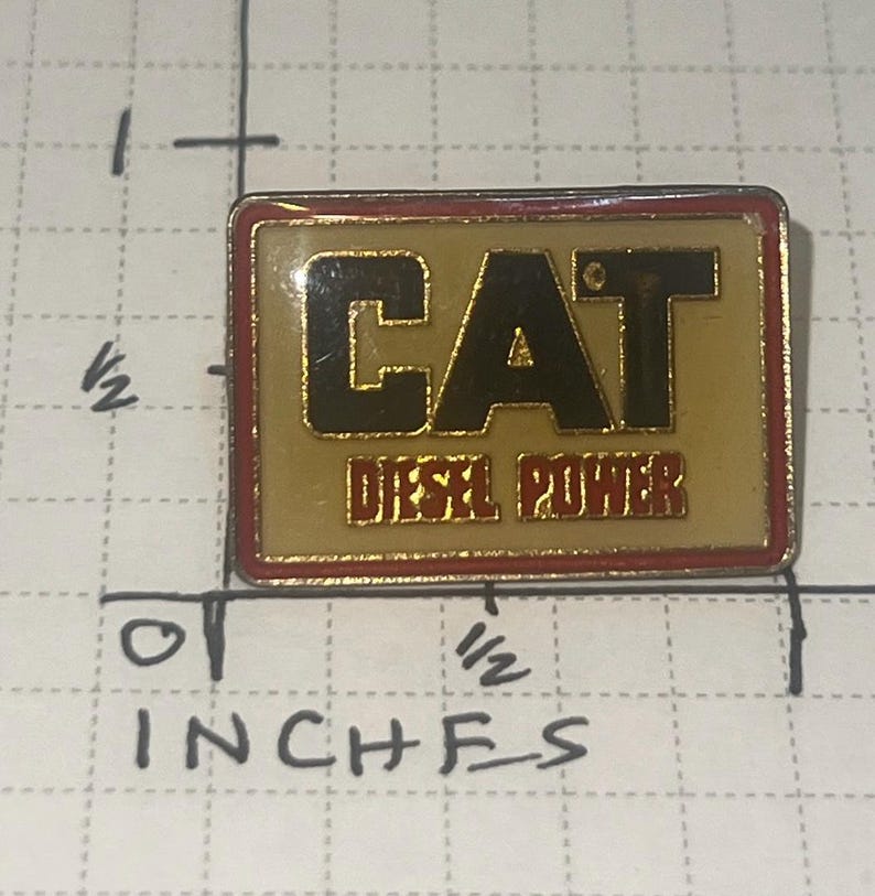 以下が含まれることがあります： 黒と赤の文字で「CAT DIESEL POWER」と書かれた長方形のピン。ピンは金色の背景に赤い縁取りがあります。ピンの長さは約1.9cm、幅は約1.3cmです。