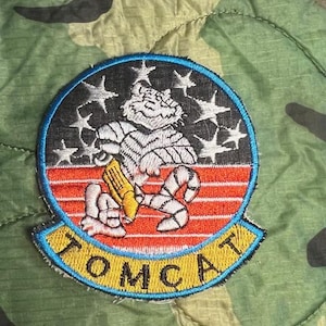 May include: A black and white embroidered patch with a cartoon tiger holding a missile. The patch has a blue border and the word "TOMCAT" in yellow letters.