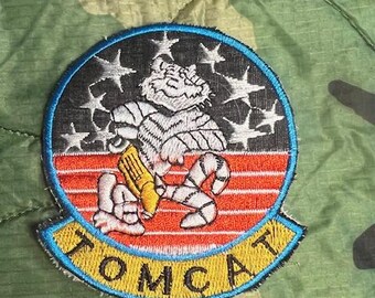 May include: A black and white embroidered patch with a cartoon tiger holding a missile. The patch has a blue border and the word "TOMCAT" in yellow letters.