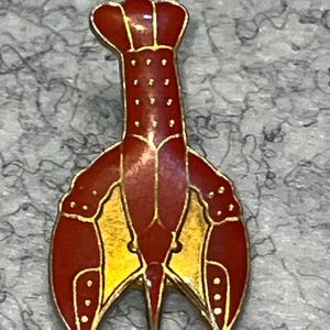 May include: A lobster-shaped enamel pin featuring a deep red body with gold-toned metal outlines and accents. The design includes a segmented tail, a central body section, and two large claws, all detailed with small gold dots against the red enamel.