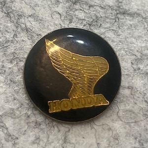 May include: A round, black Honda emblem with a gold-colored wing design and the word "HONDA" in gold lettering. The emblem has a glossy finish and is likely a pin or badge.