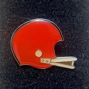 May include: A red football helmet pin with a white facemask and a gold border. The pin has a white circle on the side and is set against a dark background. A collectible item.