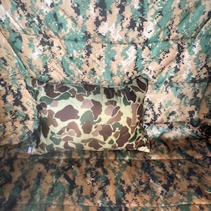 May include: A camouflage-patterned pillow with a brown, green, and tan color scheme. The pillow is sitting on a camouflage-patterned blanket.