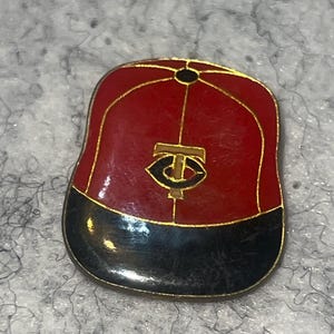 May include: A red, black, and gold baseball cap pin. The pin features the logo of the Minnesota Twins baseball team. The cap is red with a black brim and gold trim.