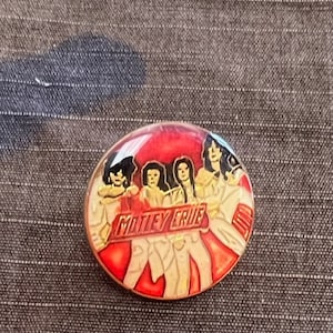 May include: A round, red and beige pin featuring the band Mötley Crüe. The pin has a red background with the band members depicted in a cartoon style. The band's name is written in beige letters.