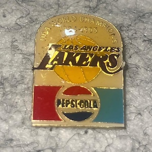 May include: A gold-colored pin featuring the Los Angeles Lakers logo with a basketball and the text "NBA WORLD CHAMPIONS 1984-1985." Below is the Pepsi-Cola logo in red, blue, and green.