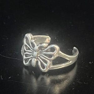 May include: A silver-colored butterfly ring. The ring features an openwork butterfly design at the center, with detailed wing patterns. The band is split, creating an adjustable fit. The ring is set against a dark background.