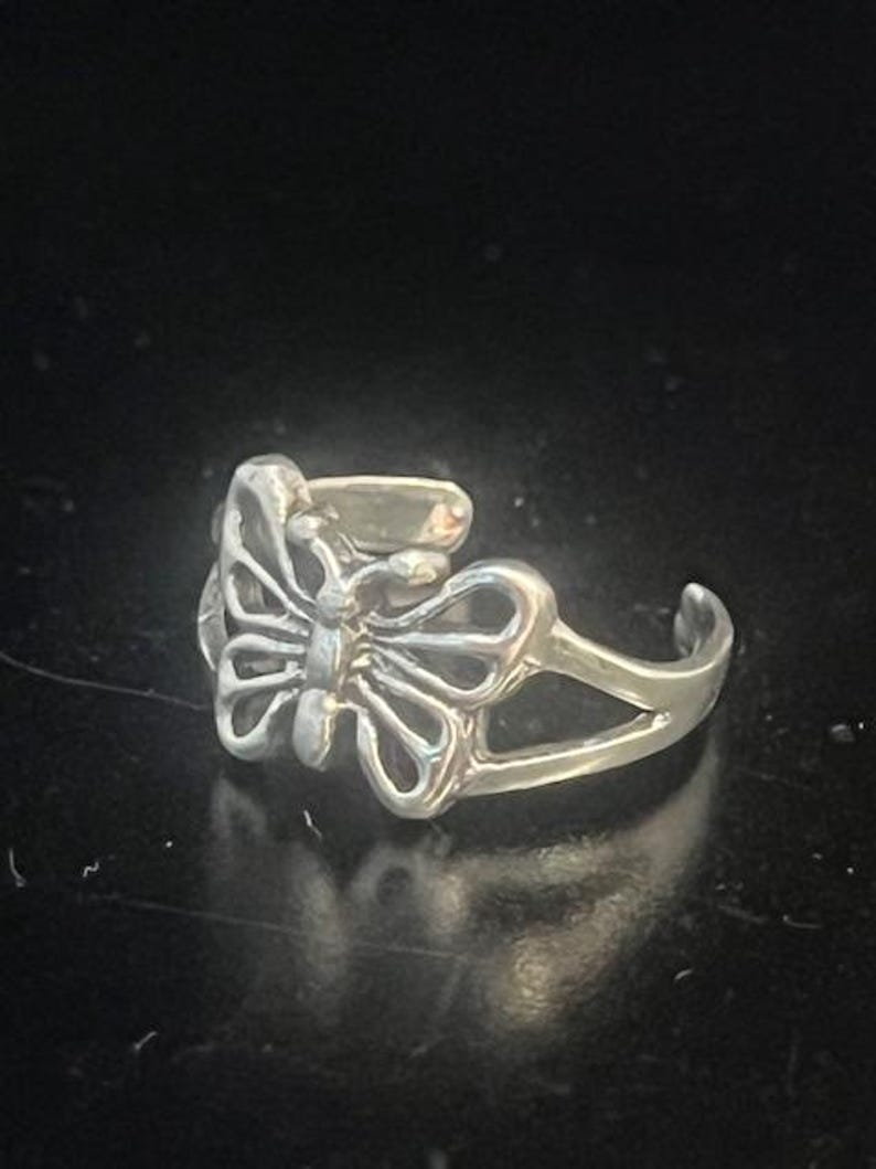 May include: A silver-colored butterfly ring. The ring features an openwork butterfly design at the center, with detailed wing patterns. The band is split, creating an adjustable fit. The ring is set against a dark background.